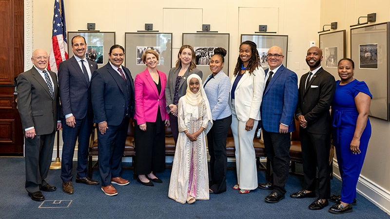 Photo of a diverse group of adults in professional attire smiling. A young girl in a white headscarf and dress stands in the middle of the adults smiling as well.