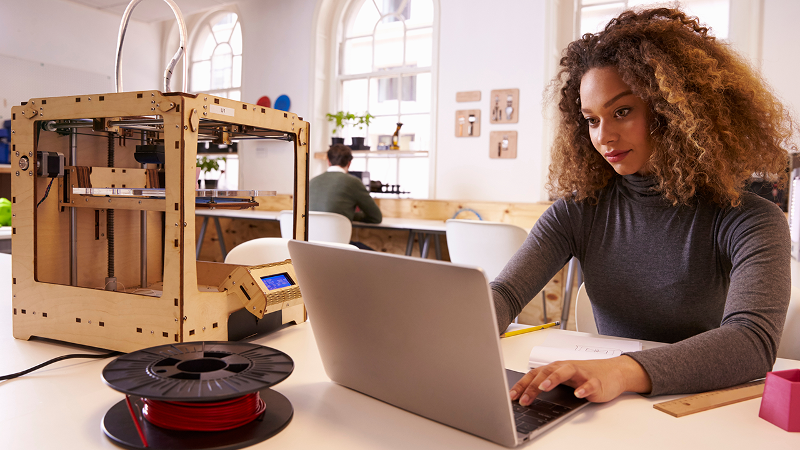 Photo of a young adult working with a laptop and 3D printer