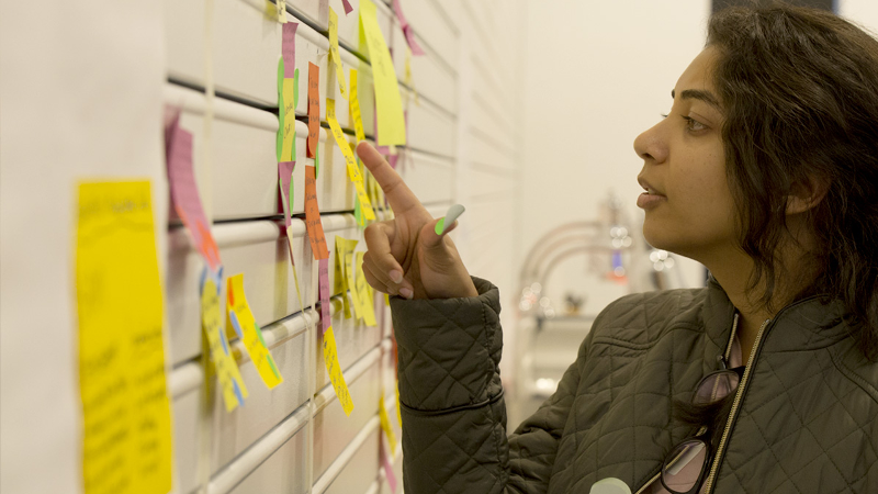 A young woman looking closely at a wall full of sticky notes and sticky dots