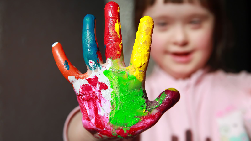 A young child with Down syndrome, showing her five fingers painted colorfully