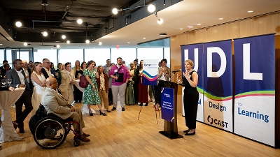 Photo of CAST CEO Lindsay Jones speaking to event guests, standing in front of three banners spelling out UDL.