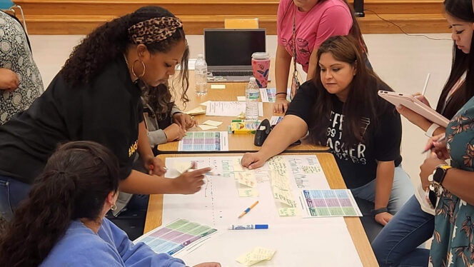 Team of educators working together at a long table covered with markers, guidelines, and paper.
