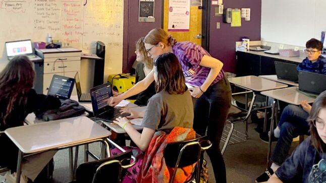 Students are using laptops at their desks, a teacher leans over to point to one of the screens.