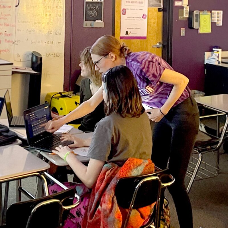 Students are using laptops at their desks, a teacher leans over to point to one of the screens.