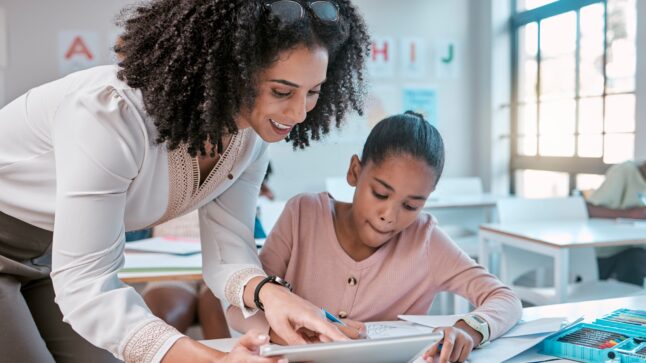 Teacher and young learner looking at a tablet - the learner is sitting at her desk in a classroom with her materials and supplies at the ready.