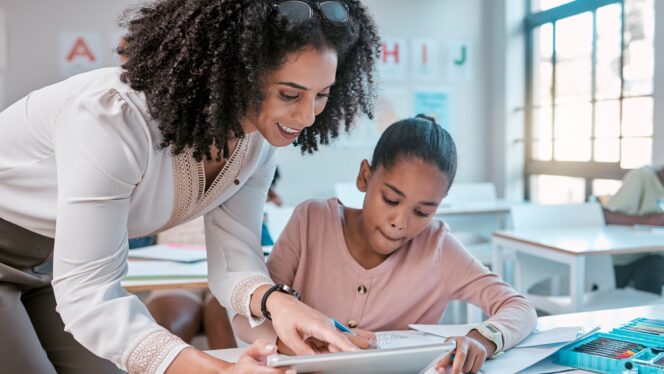 Teacher and young learner looking at a tablet - the learner is sitting at her desk in a classroom with her materials and supplies at the ready.