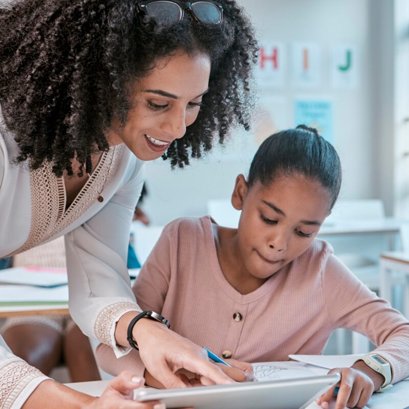 Teacher and young learner looking at a tablet - the learner is sitting at her desk in a classroom with her materials and supplies at the ready.