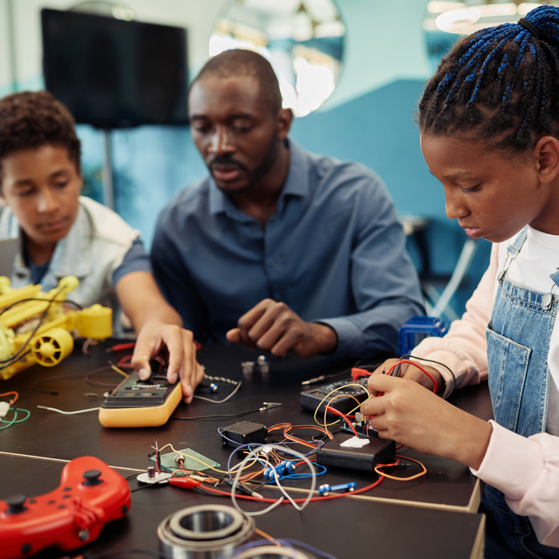 Teacher working with two students on a robotics lab project at a table with electronics components and tools.