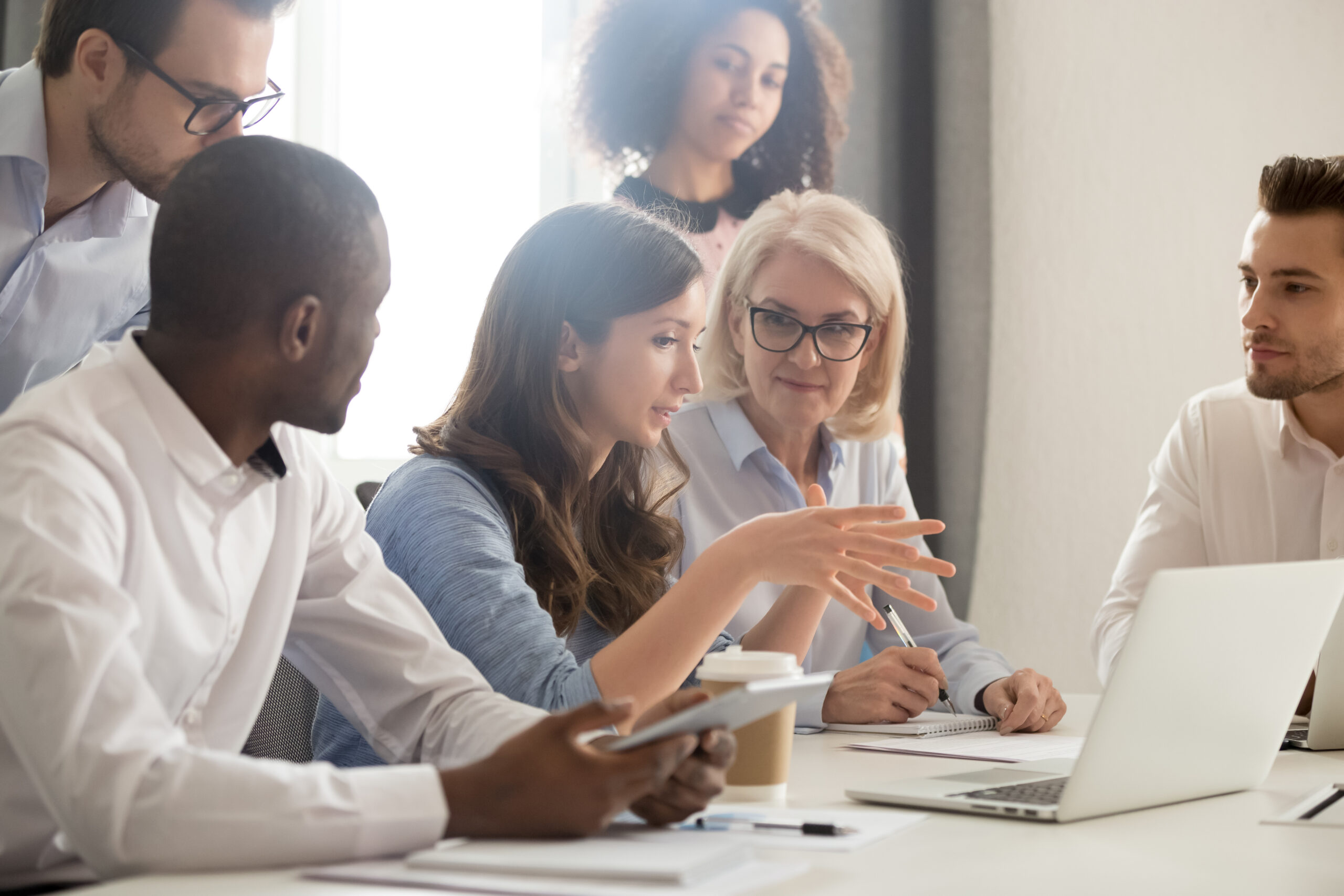 Young female mentor leader coach teaching employees group analyzing online project explaining business strategy speaking training diverse corporate team with laptop using computer at office meeting.