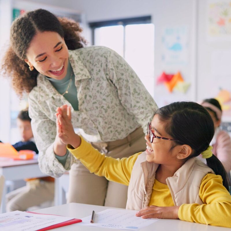 Teacher woman, high five girl and classroom with achievement, success and mentorship for learning. Education, development and students with goals, knowledge and books with celebration at school desk