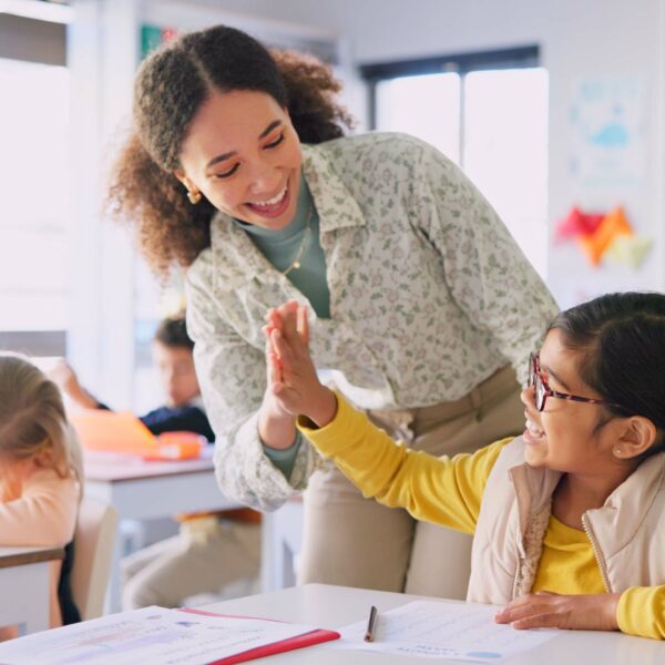 Teacher woman, high five girl and classroom with achievement, success and mentorship for learning. Education, development and students with goals, knowledge and books with celebration at school desk.