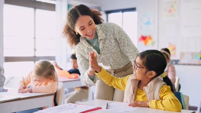 Teacher woman, high five girl and classroom with achievement, success and mentorship for learning. Education, development and students with goals, knowledge and books with celebration at school desk.