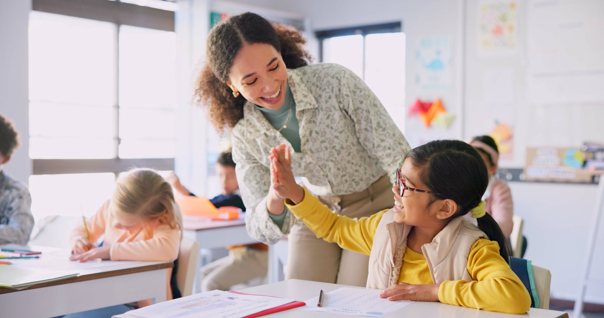 Teacher woman, high five girl and classroom with achievement, success and mentorship for learning. Education, development and students with goals, knowledge and books with celebration at school desk.