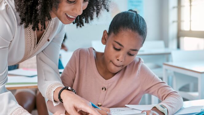 Female teacher helping young girl, teaching creative work in book and children in sunny classroom. Education empowerment in school, educator speaking to student and learning mathematics at desk