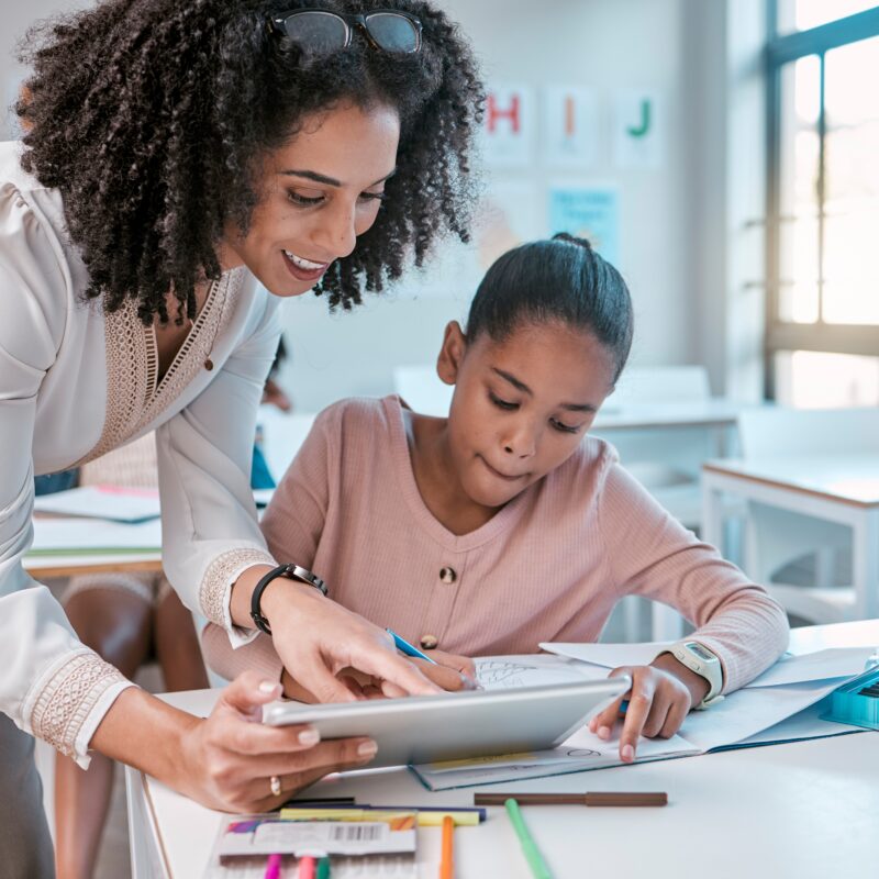 Female teacher helping young girl, teaching creative work in book and children in sunny classroom. Education empowerment in school, educator speaking to student and learning mathematics at desk