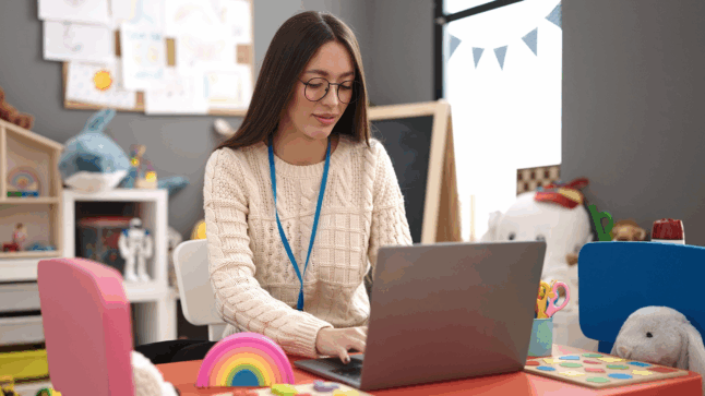 Photo of a teacher using a laptop at her desk in a colorful kindergarten classroom.