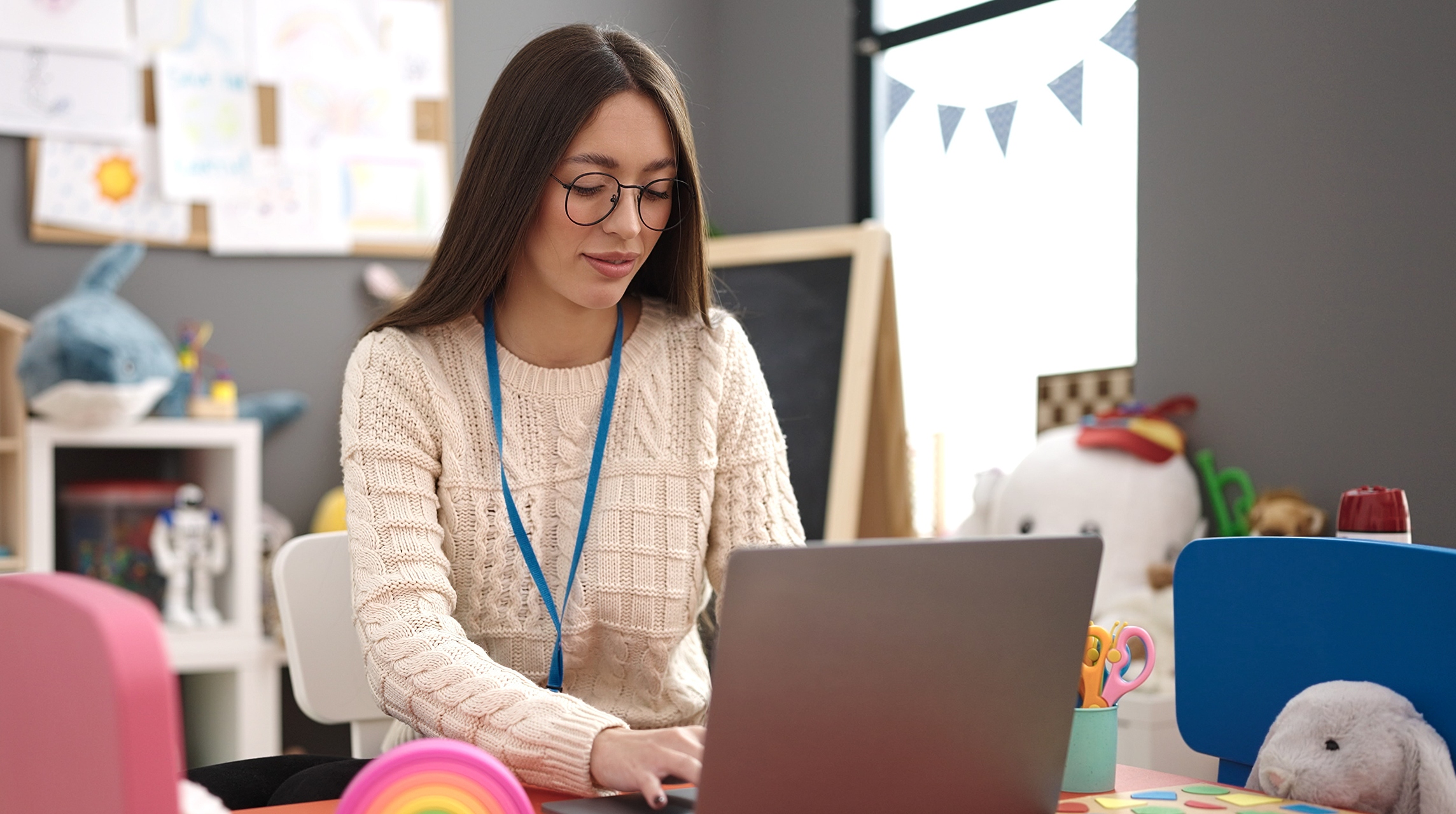 Photo of a teacher using a laptop at her desk in a colorful kindergarten classroom.