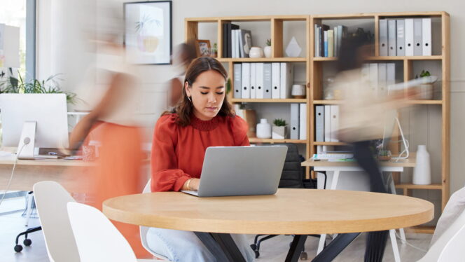 an office scene where a woman sits at a table at her laptop while blurry co-workers zip past around her in the background
