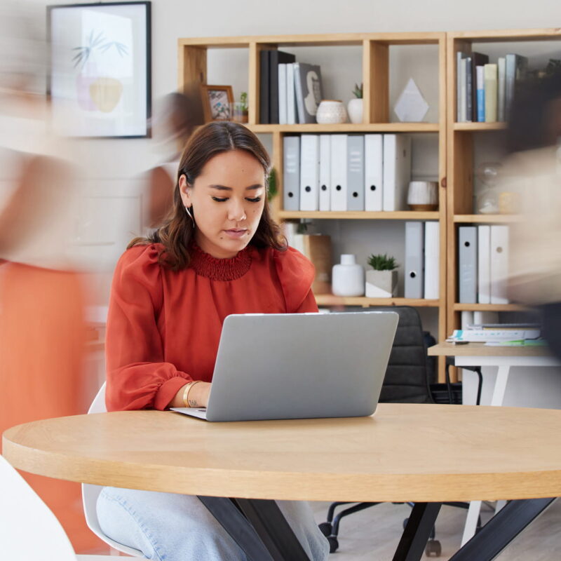 an office scene where a woman sits at a table at her laptop while blurry co-workers zip past around her in the background