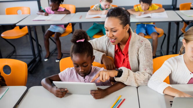 Students seated at their tables working independently while one student has the teacher leaning over them smiling as they use a tablet