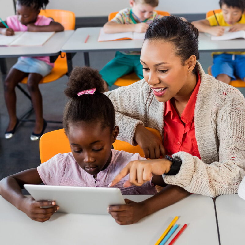 Students seated at their tables working independently while one student has the teacher leaning over them smiling as they use a tablet